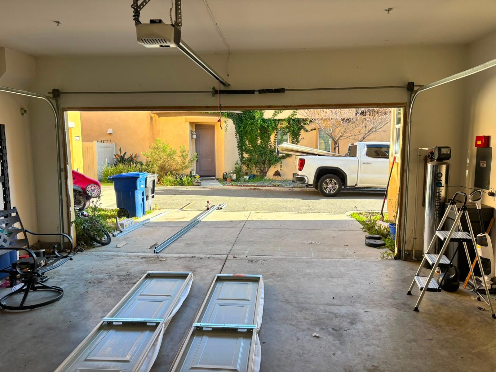 A garage door part way through repair, two panels removed and standing on the floor, with the opener and a broken spring visible above the open doorway