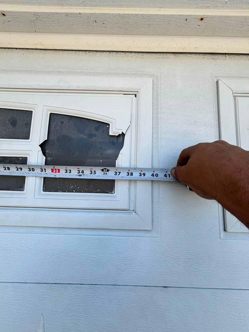 A tape measure held across a white garage door panel with a broken decorative window insert, measuring the opening before ordering a replacement