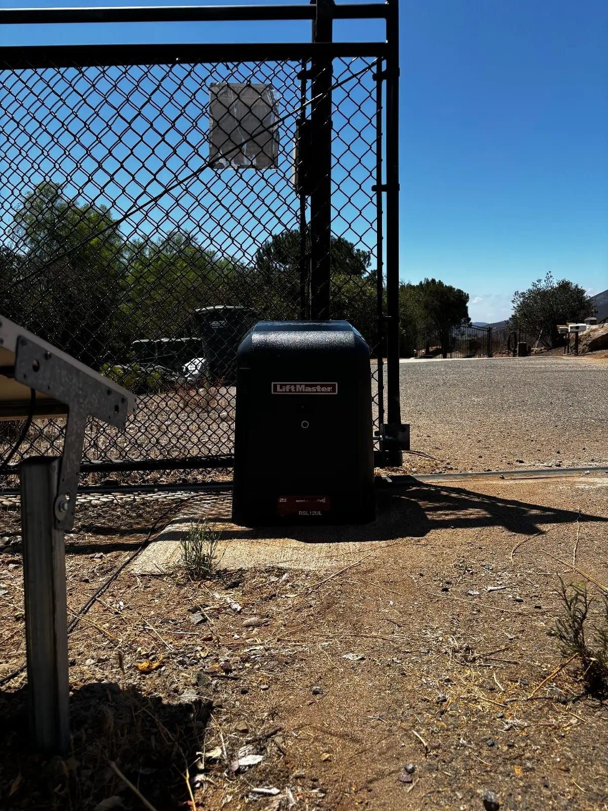A black LiftMaster slide gate operator on a concrete pad beside a chain link driveway gate, with the gate control box mounted on the frame above it